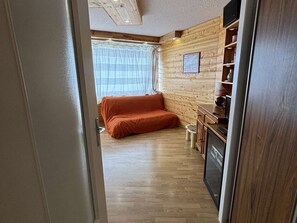 A compact room with a wooden floor, a bed with an orange blanket, a small kitchenette with a stove, and a window with sheer curtains. - Cozy apartment in Alpe d'Huez - 5 guests (Huez)