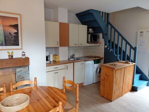 A compact kitchen with white cabinets, a microwave, and a washing machine. There's a wooden table with chairs and a basket. A staircase with blue railings leads to another level. - Cozy duplex apartment with view - 6 guests (Esquièze-Sère)