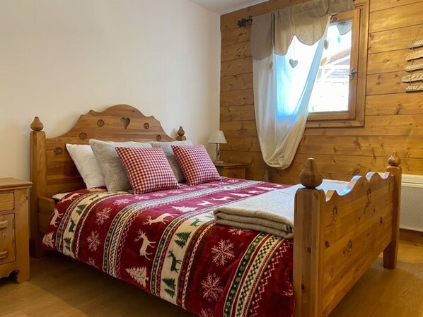 Uma cama de madeira com travesseiro xadrez, manta estampada e janela com cortinas.