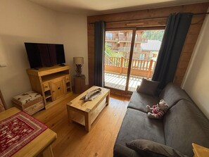 A cozy living room with a sofa, a coffee table, a TV on a wooden stand, and a view of a wooden deck through a glass door. - T3 Apartment with WIFI - 4/5 guests, south balcony (Crest-Voland)
