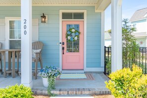A house with a pink front door, a number '200' on the wall, and a porch with chairs and a potted plant. - Beau Coast Retreat | Pool + Near Waterfront (Beaufort)