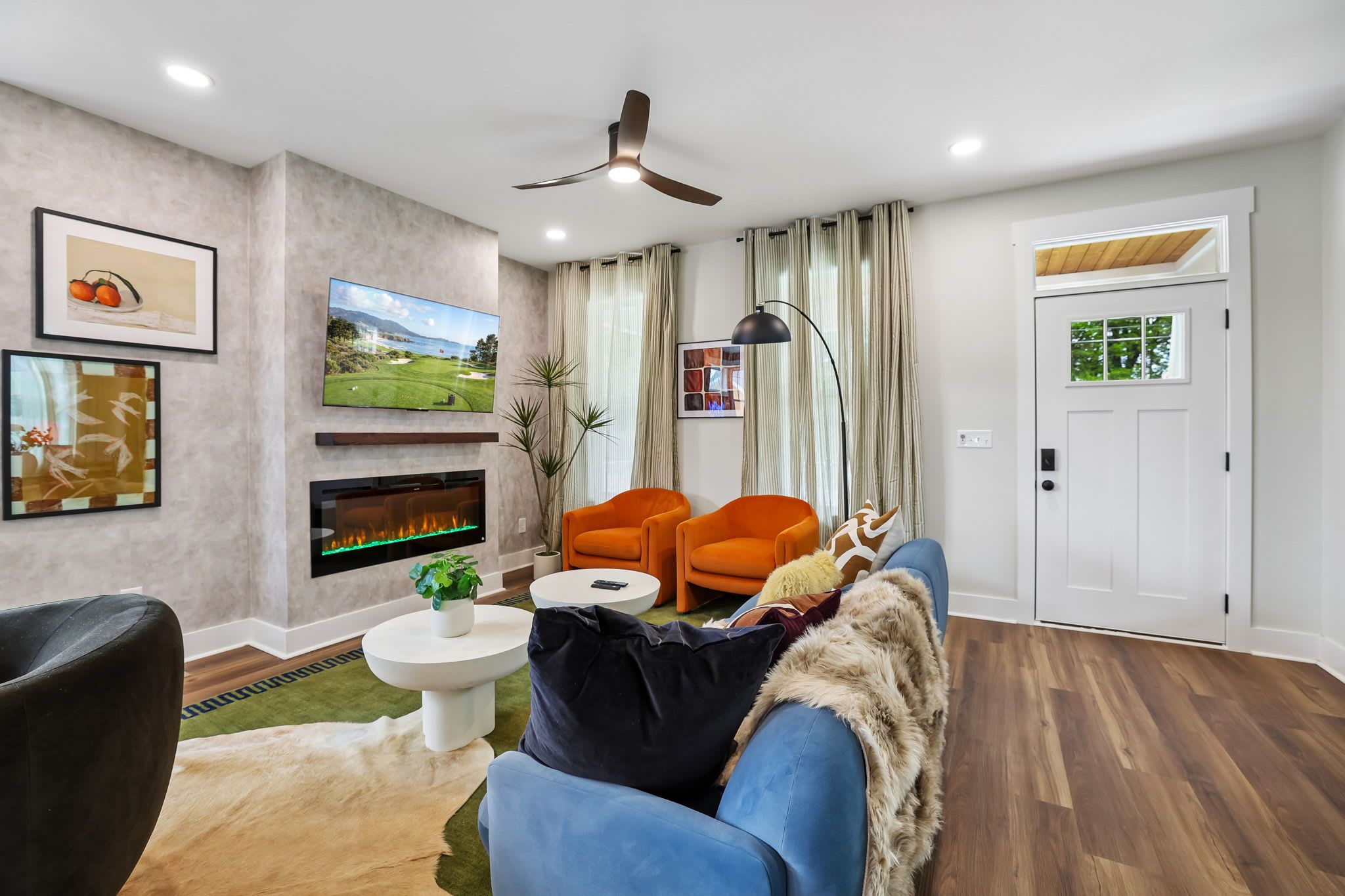 A modern living room with a flat-screen TV, a fireplace, two orange armchairs, a blue sofa, and a ceiling fan.