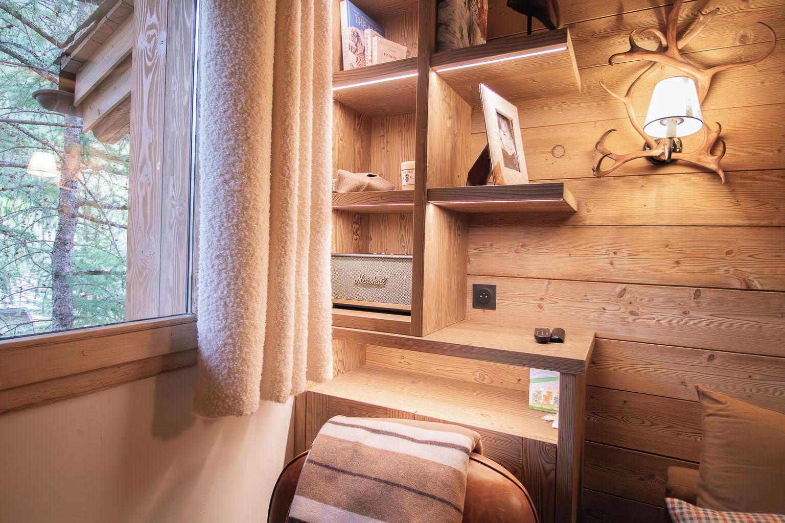 Interior acogedor de una cabaña con paredes de madera, una ventana con cortinas, una estantería con libros, una lámpara y un banco acolchado.