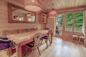 A rustic wooden dining area with a bench, chairs, and a table. There are two hanging light fixtures and a large window with a view of greenery. - Roche des Fours 210 (Val-d'Isère)