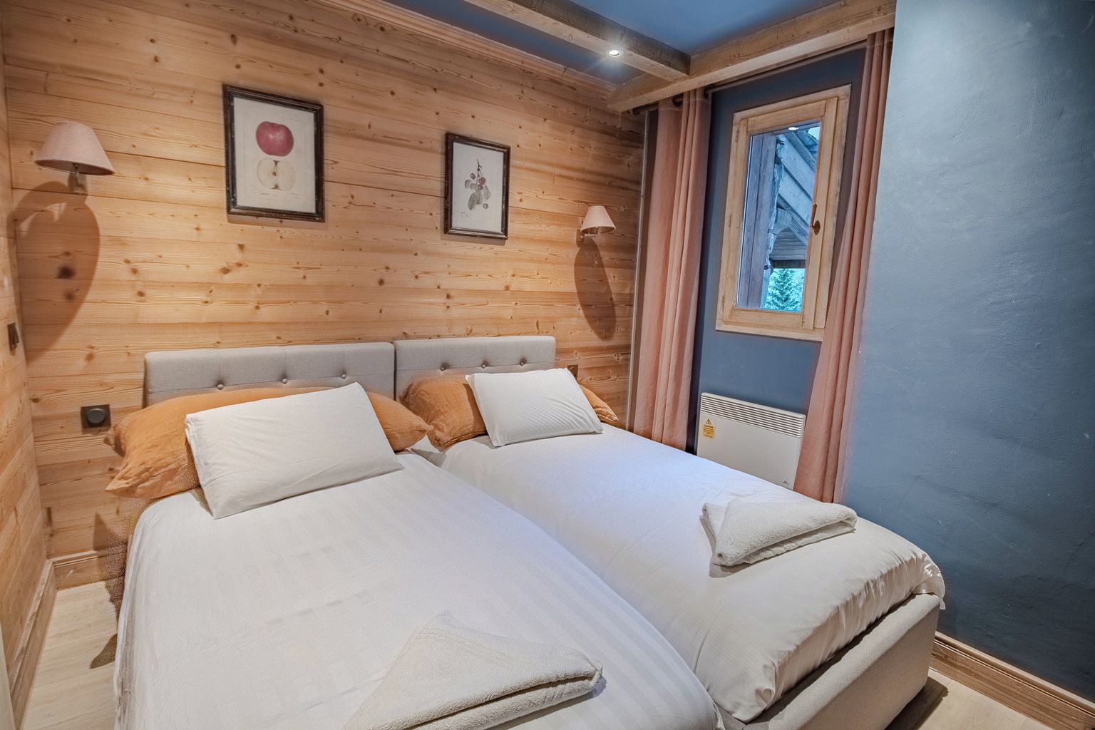 Una cama doble con dos almohadas y una colcha, paredes de paneles de madera, una ventana con cortinas y lámparas fijadas a la pared.