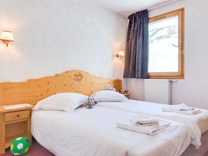 A hotel room with a wooden headboard, two beds, a nightstand, a lamp, a window with curtains, and a soccer ball on the floor. - Spacious apartment - 6 guests, ski-in/ski-out (Les Menuires)