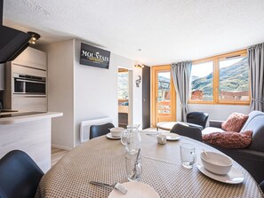 A modern kitchen with a built-in oven, a dining area with a round table set for two, and a sofa with a fluffy pillow. - Comfortable apartment with mountain view - 4 guests (Les Menuires)