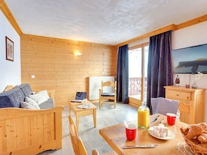 Ein gemütliches Zimmer mit Holzboden, einer Couch, einem Esstisch mit Frühstücksgebäck und einem Laptop, sowie einem Blick durch das Fenster.