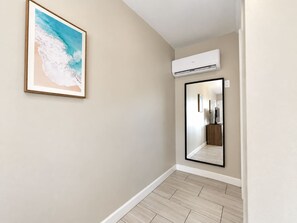 A hallway with a framed artwork, an air conditioning unit, and a mirror reflecting a room. - Coastal Daydream at 300 Tenth | 103 (Myrtle Beach)
