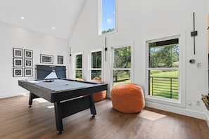 A modern indoor space with a pool table, bean bag seating, and large windows offering views of a green landscape. - The Treehouse, Sleeps 12, Waterfront, Game Room, Firepit, Kayaks + MORE! (Milledgeville)