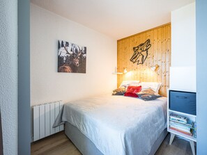 A bedroom with a bed, pillows, a bedside table with books, a wall-mounted picture, and a wooden headboard. - Pleasant apartment, near slopes - 4 guests (Les Menuires)