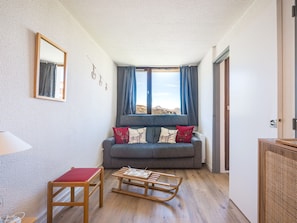 A cozy living room with a grey sofa, a wooden rocking chair, and a view of the outdoors through a window with blue curtains. - Pleasant apartment, near slopes - 4 guests (Les Menuires)