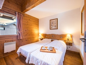 A wooden-paneled room with two beds, a window with curtains, a wall-mounted picture, and a bedside table with a lamp. - Spacious apartment - 6 guests (Les Menuires)