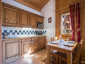 A wooden kitchen with a tiled backsplash, a dining table set for two, and a window with curtains. - Spacious apartment - 6 guests (Les Menuires)