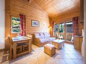 A wooden cabin interior with a dining table, sofa, and bench, a picture on the wall, and a view through the window. - Spacious apartment - 6 guests (Les Menuires)
