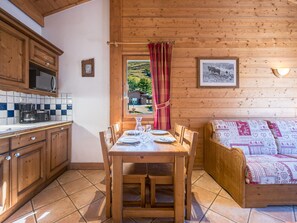A cozy cabin interior with a dining table set for four, wooden cabinets, a kitchen area, and a window with a view of a rural landscape. - Spacious apartment - 6 guests (Les Menuires)