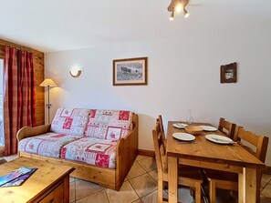 Een gezellige kamer met een houten bank, een eettafel met stoelen, een lamp en lijstjes met foto’s aan de muur.