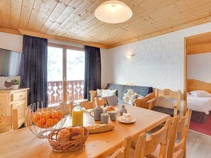 A room with a wooden dining table set with breakfast items, a bed with a wooden headboard, and a window with curtains. - Spacious ski-in/ski-out apartment - 8 guests (Les Menuires)