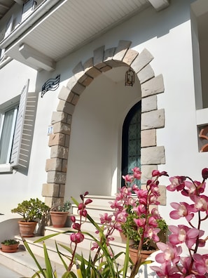Ein Gebäude mit heller Fassade, einem steinernen Bogen und einem Fenster mit Metallgitter. Vor dem Eingang stehen Pflanzgefäße.