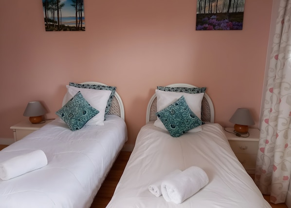 Ein Hotelzimmer mit zwei Betten, jeweils mit einer weißen Decke, einem aufgerollten weißen Handtuch und gemusterten Kissen. Auf den Nachttischen stehen jeweils zwei Nachttischlampen.
