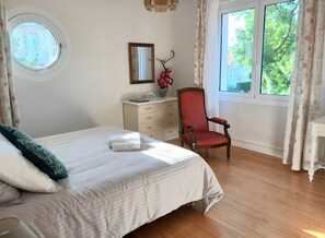 Ein Schlafzimmer mit Bett, Stuhl, Kommode, Spiegel und einem Fenster mit Vorhängen.