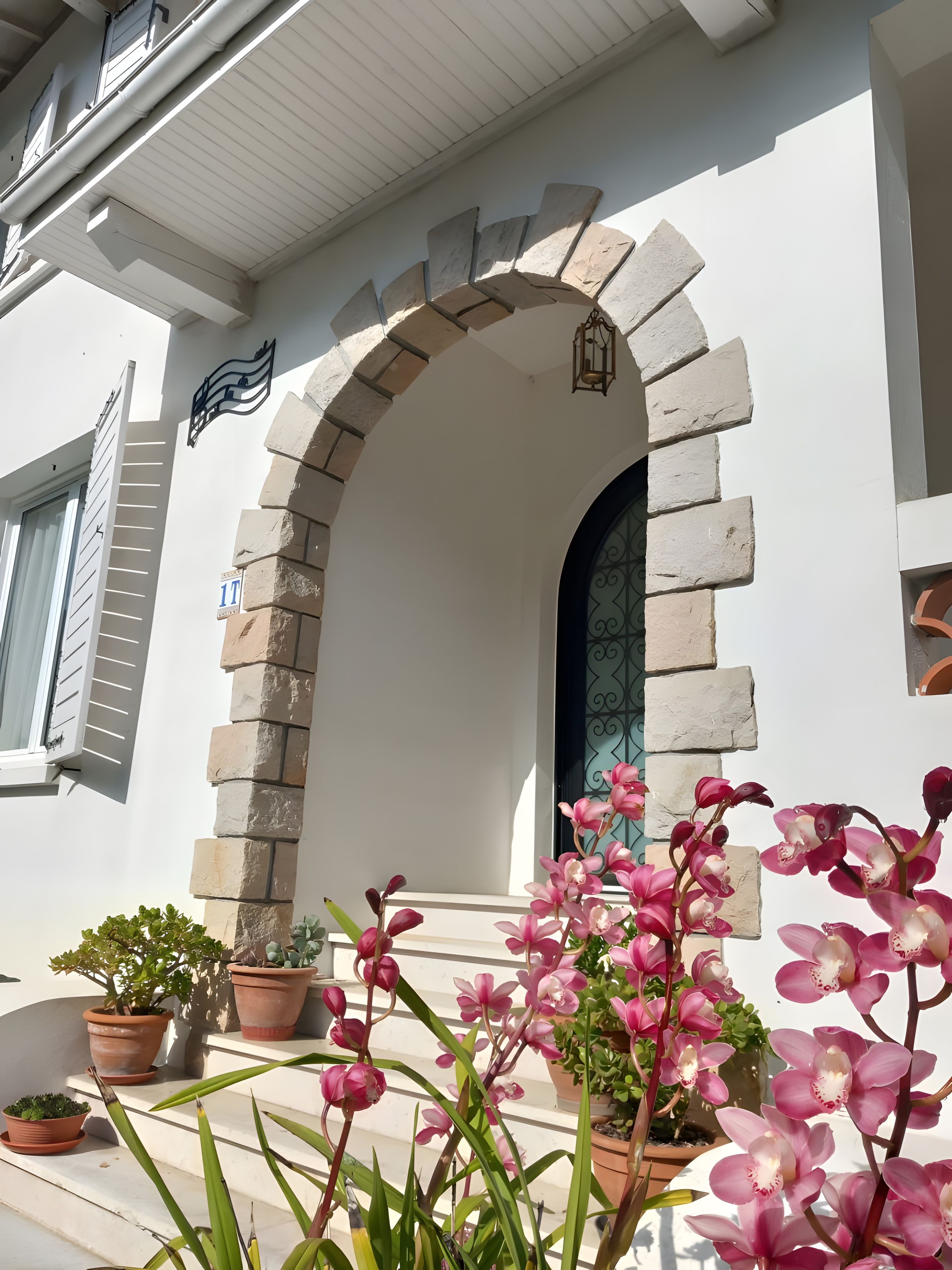 Un bâtiment à la façade blanche, doté d’une arche en pierre et d’une fenêtre ornée d’une grille métallique. Des plantes en pot sont disposées devant l’entrée.