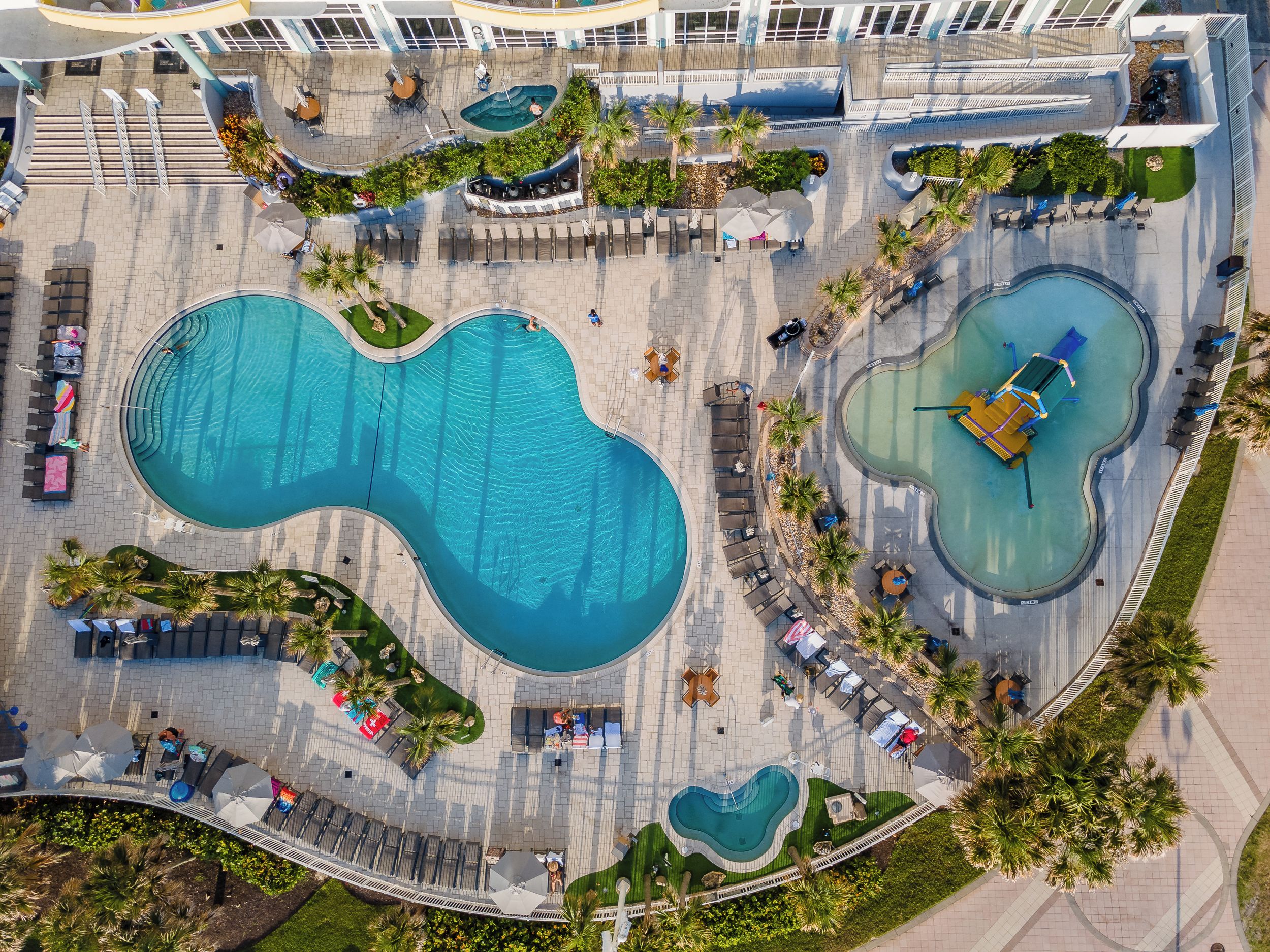 A large outdoor swimming pool with multiple lanes, surrounded by lounge areas and palm trees.
