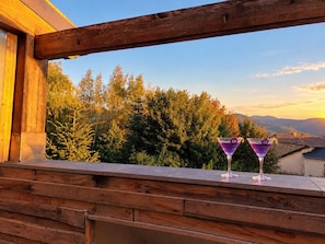 A wooden balcony with two martini glasses and a view of trees and mountains.