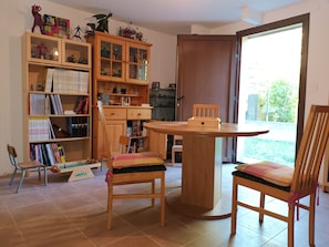 Une pièce avec un meuble en bois rempli de livres et de jouets, une table ronde en bois entourée de chaises, et une vue sur l’extérieur par une porte.