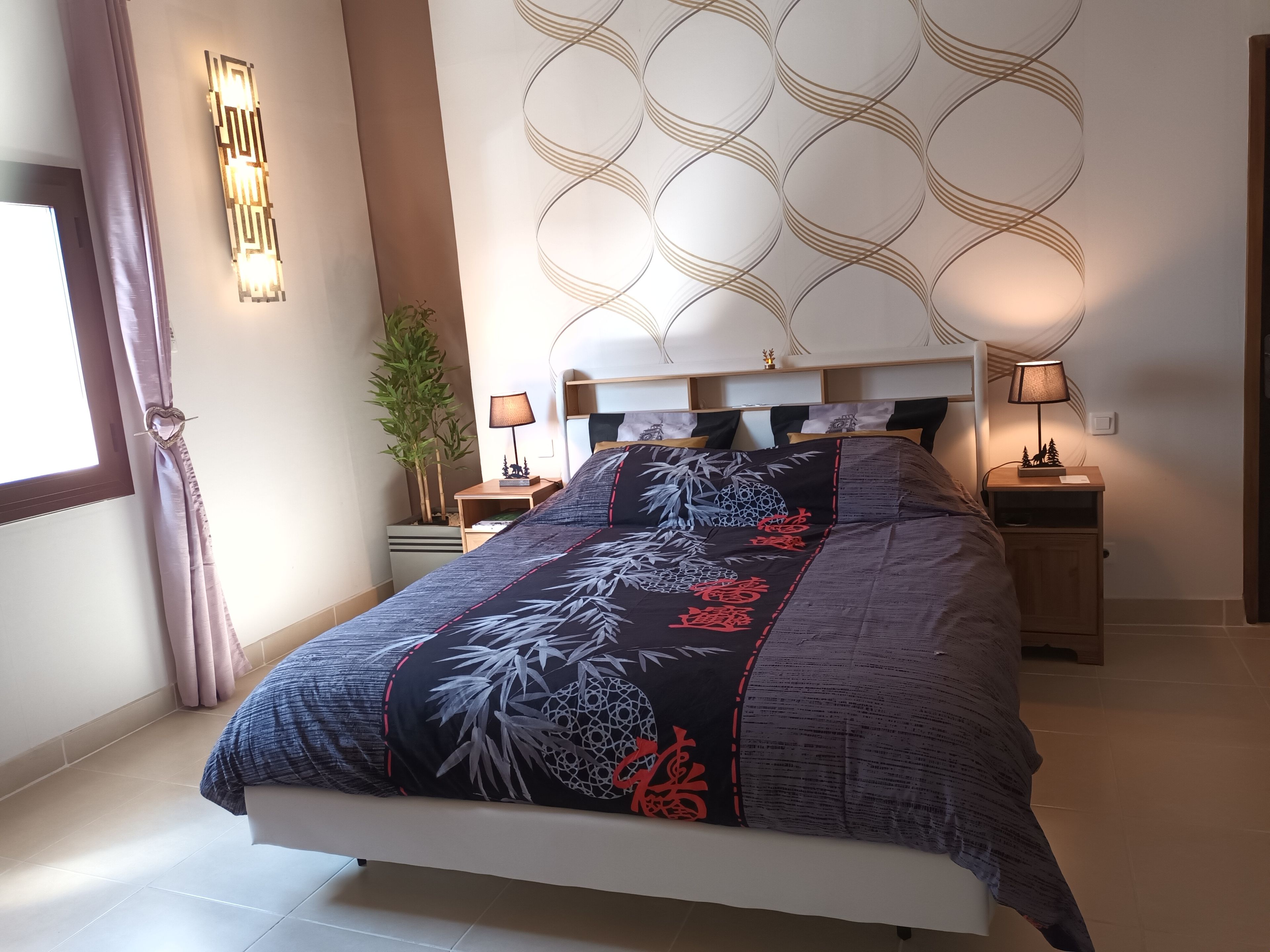 A modern bedroom with a large bed, bedside tables, a wall-mounted light fixture, and a potted plant.