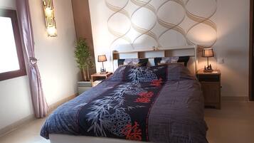 A modern bedroom with a large bed, bedside tables, a wall-mounted light fixture, and a potted plant.