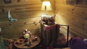 A cozy cabin interior with wooden walls, a small table with various items, a lamp, and a chair.