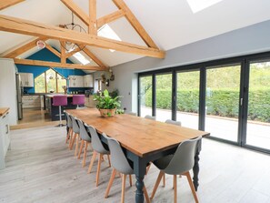 A spacious dining area with a long wooden table and modern chairs, a kitchen with blue accents, and large windows offering a view of the outdoors. - Hazeldene (Evesham)
