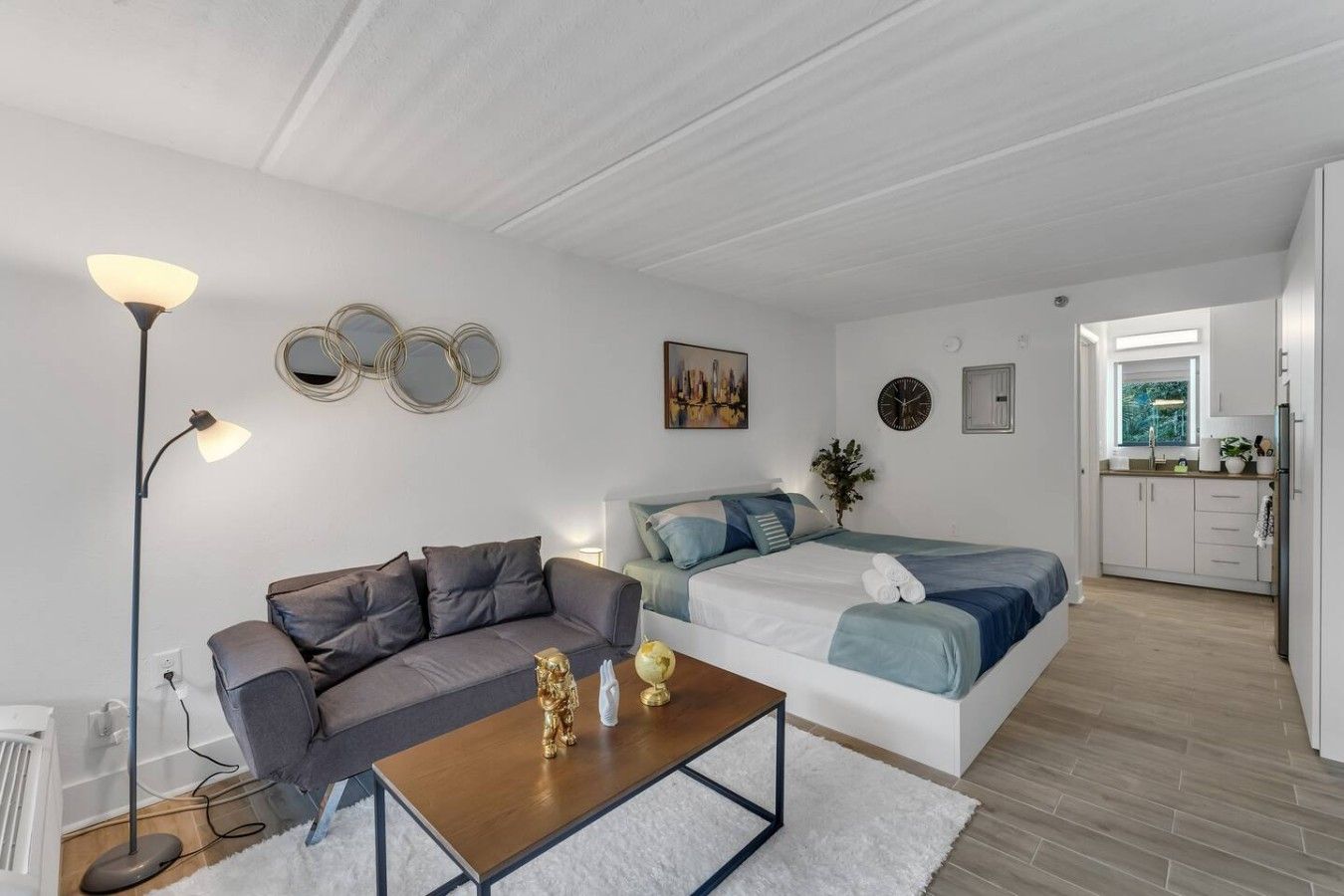 Habitación moderna y bien iluminada con sofá, cama, una mesita de centro y una cocina visible a través de un umbral abierto.