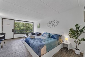 A bedroom with a bed, a desk, a chair, a potted plant, and a view of trees through a window. - Orlando Studio | Disney Nearby + Resort Pool | Orl (Kissimmee)