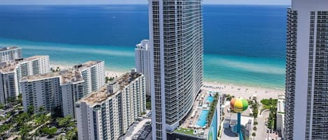 A high-rise hotel with a pool and beach access, surrounded by other buildings and a clear view of the ocean.