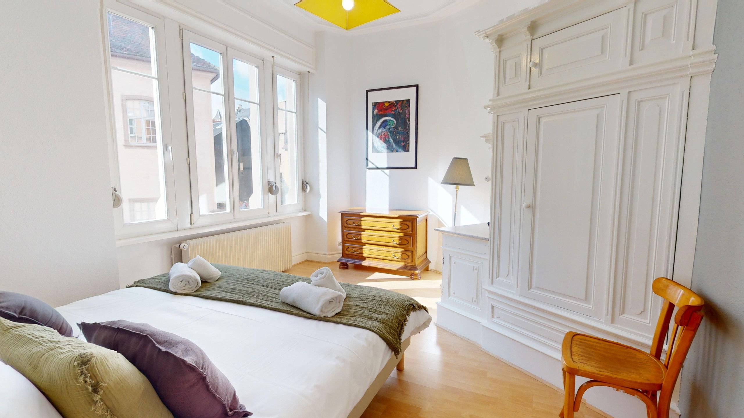 A hotel room with a bed, a wooden dresser, a chair, and a large window.