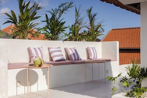 A sunlit outdoor seating area with a white wall, striped cushions, and a coconut drink on a small round table.
