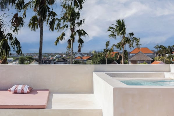 A pool area with a lounge chair and a cushion, surrounded by palm trees and buildings in the background.