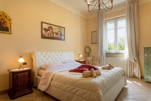 A bedroom with a large bed, a nightstand, a dresser, a window with curtains, and a chandelier. - Holiday home 'Antico Borgo Lucca - Pool And' with Wi-Fi and air conditioning (Lucca)