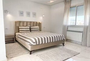 A bedroom with a bed, nightstands, a window with curtains, and wall-mounted lights. - The Dream House (Marsa)