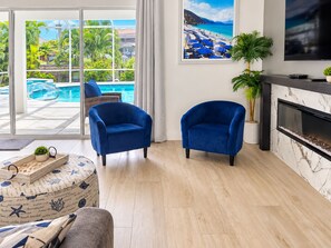 A modern living room with a pool view, two blue armchairs, a fireplace, and a large TV on the wall. - Cape Coral Waterfront Retreat with Heated Pool & Dock (Cape Coral)