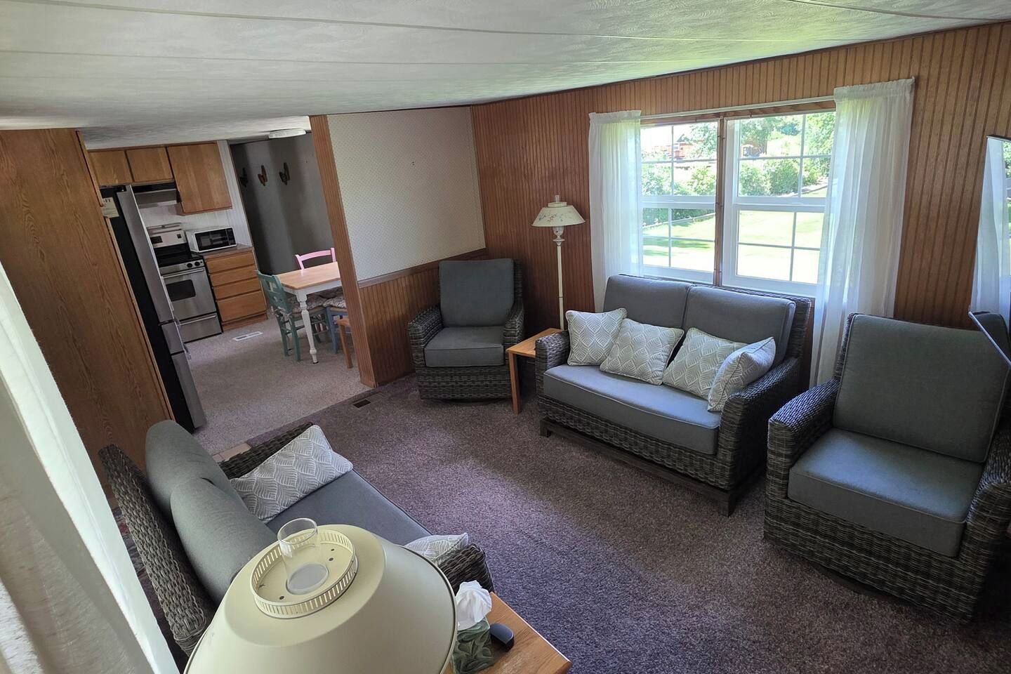 A compact living space with a sofa, two armchairs, a small table, and a lamp. A kitchenette with a stove and refrigerator is visible in the background.