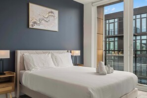 A hotel room with a large bed, two bedside lamps, a painting on the wall, and a view of the city through a large window. - Urban 1BR Escape | Northern Liberties (Philadelphia)