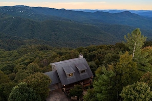 A house with a sloped roof, surrounded by dense forest and mountains in the background. - Mtn Views, Game Room, Fire Table, Porches (Blowing Rock)