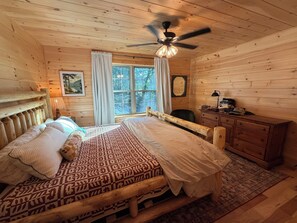 A wooden cabin bedroom with a bed, a desk, a chair, a ceiling fan, and a window. - The Friendly Fern - Welcoming Old Friends and New (Old Forge)