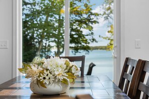 A white vase with flowers on a wooden table, a wooden chair, and a view of trees and water outside. - Sleeping Bear Dunes Lakefront Getaway (Empire)