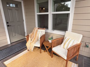 Eine Veranda mit zwei Holzstühlen, einem kleinen Tisch, einem Blumentopf und einer Tür mit Fußmatte.