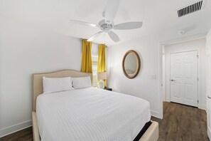 A bedroom with a bed, a ceiling fan, a window with yellow curtains, a round mirror, and a white door. - The Big Cat 3BR | Lanai | Near Brownwood (The Villages)