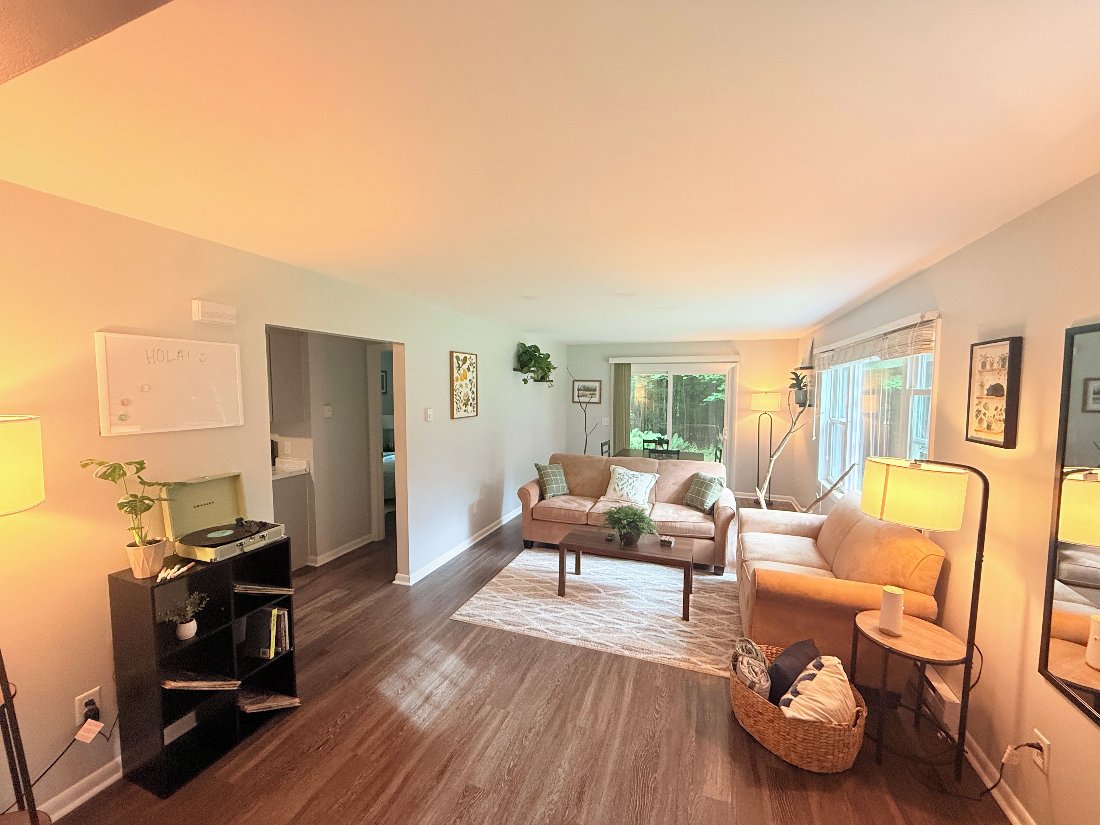 A living room with a sofa, a coffee table, and a lamp. There is a bookshelf and a plant on the left side.